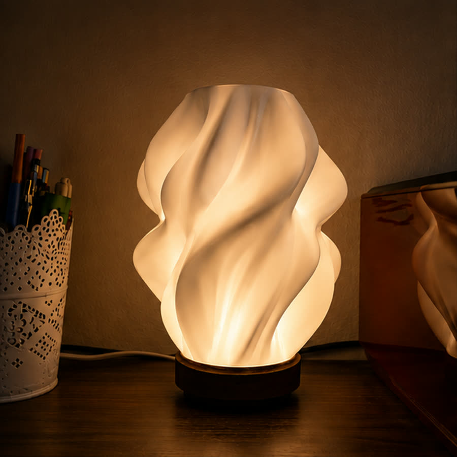 The Sculptural Wave Table Lamp lit on a wooden desk beside a pen pot